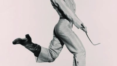 A person dressed in traditional attire and hat leaps energetically in mid-air while holding a stick, against a plain background. [gen-ai]