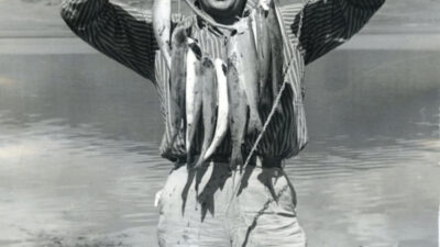 A man stands by a lake holding up a string of five fish, with a small rowboat and mountains visible in the background. [gen-ai]
