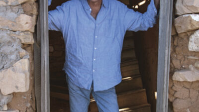 A man in a light blue shirt and jeans stands smiling in a stone doorway of a rustic, partially collapsed building. [gen-ai]