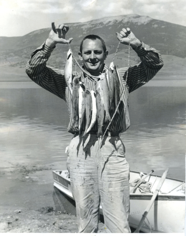 A man stands by a lake holding up a string of five fish, with a small rowboat and mountains visible in the background. [gen-ai]