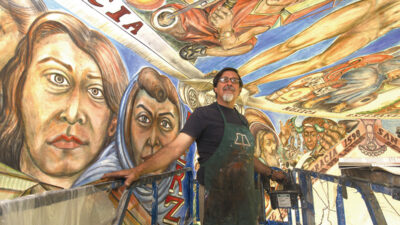 An artist stands on a lift, holding the railing, in front of a colorful mural with large, expressive faces and figures painted on the wall and ceiling. [gen-ai]
