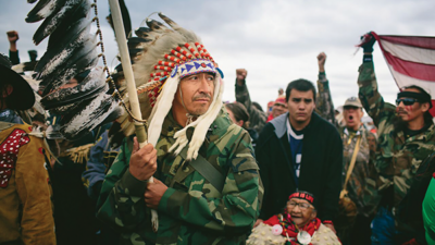 A man in a feathered headdress and camouflage jacket stands among a group of people, some raising fists, with a cloudy sky in the background. [gen-ai]