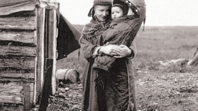 A woman wearing traditional clothing stands outside a wooden structure, holding a small child dressed warmly; both face the camera in a rural setting. [gen-ai]