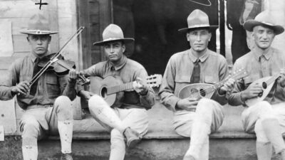 Four men in uniform and hats sit on steps, playing string instruments including a violin, guitar, mandolin, and banjo, in front of a wooden building. [gen-ai]