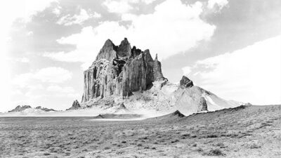 Black-and-white photo of a large, jagged rock formation rising from a flat, sparsely vegetated plain under a partly cloudy sky. [gen-ai]