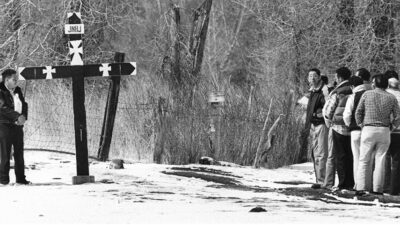 A group of people stands and kneels near a roadside memorial with large crosses in a snowy, rural area bordered by trees and a wire fence. [gen-ai]