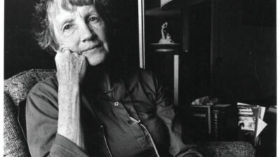 An older woman with short hair sits in an armchair, resting her head on her hand, with several books stacked on her lap. A lamp and bookshelf are visible in the background. [gen-ai]