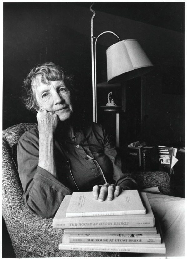 An older woman with short hair sits in an armchair, resting her head on her hand, with several books stacked on her lap. A lamp and bookshelf are visible in the background. [gen-ai]