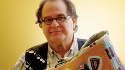 A person with round glasses and dark hair stands in front of a yellow background, holding a colorful woven basket featuring geometric patterns. [gen-ai]