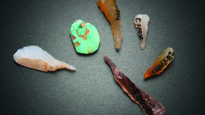 Six assorted stone artifacts, including various arrowhead and tool shapes, are arranged on a gray surface; some have labels or numbers written on them. [gen-ai]