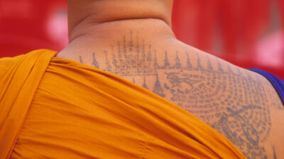 A Buddhist monk with a shaved head displays intricate tattoos on the scalp and upper back, wearing an orange robe, standing before a red background. [gen-ai]