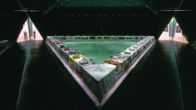 A large triangular table set with ornate place settings and decorated plates, displayed in a spacious, dimly lit room with dark walls and spotlights. [gen-ai]