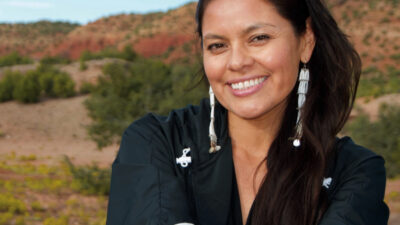 A woman with long dark hair, wearing a black jacket and beaded earrings, smiles outdoors in a desert landscape with hills in the background. [gen-ai]