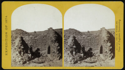 Stereograph of a partial stone ruin in a desert landscape, with two people seated beside the structure. Text on border reads Expedition of 1874 and credits Lt. Geo. M. Wheeler. [gen-ai]