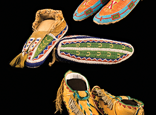 Three pairs of Native American moccasins with detailed beadwork and leather fringe are displayed against a black background. [gen-ai]