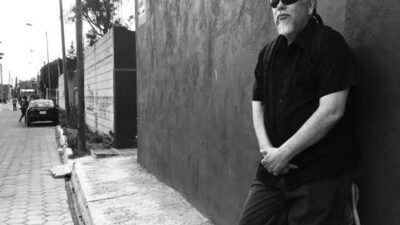 A man with a beard and sunglasses leans against a wall on a sidewalk in an urban area, with cars and trees in the background. The image is in black and white. [gen-ai]