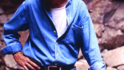 A man in a denim shirt and jeans stands outdoors on rocky terrain, resting one hand on his hip and the other on his knee, looking to his left. [gen-ai]