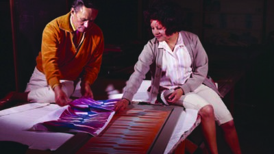Two people sit at a table examining and discussing colorful abstract artwork illuminated by overhead light in a dim room. [gen-ai]