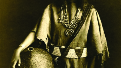 A Native American woman in traditional clothing stands holding a large vessel, wearing beaded jewelry and a decorated belt, looking at the camera. [gen-ai]