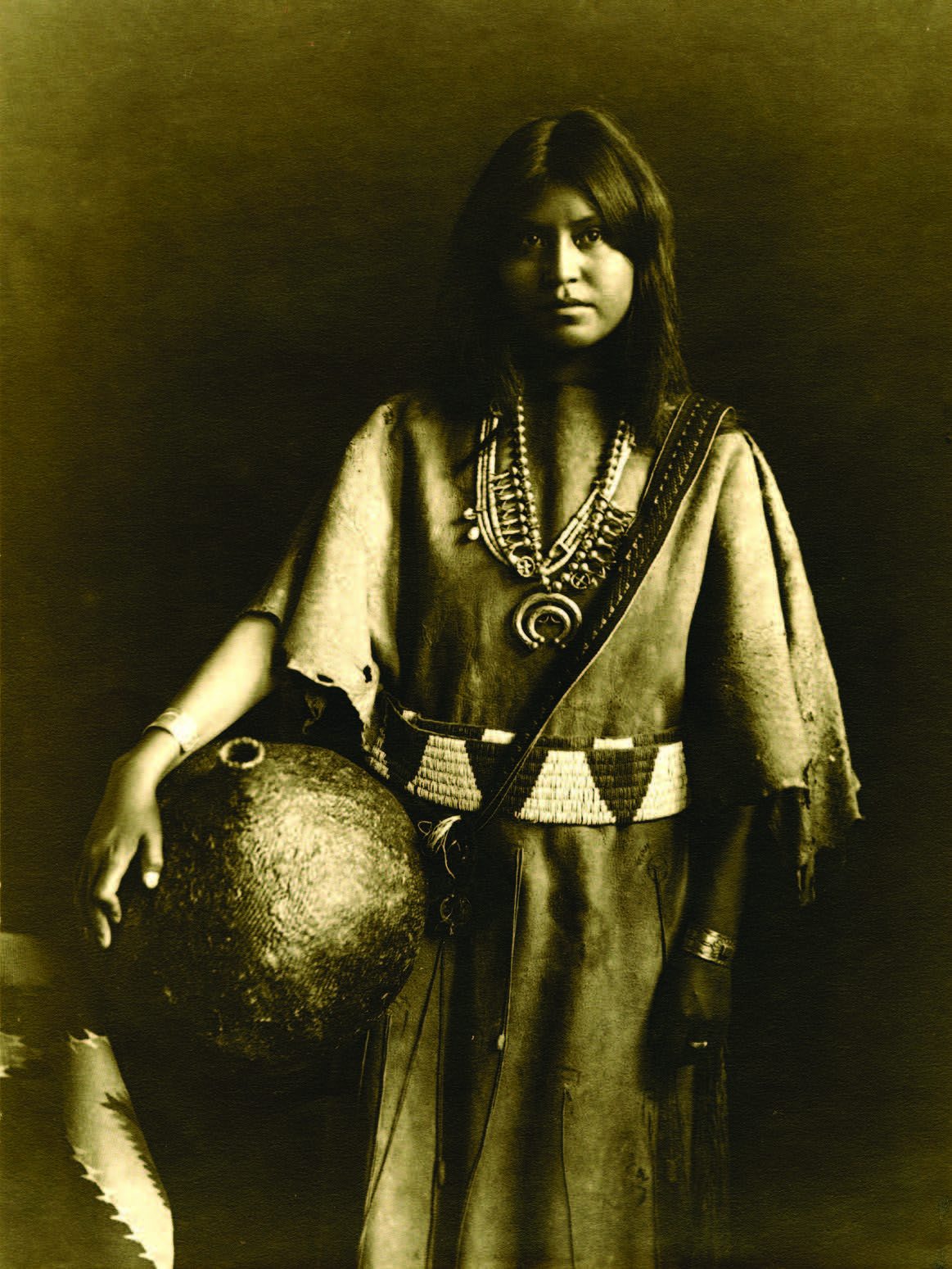 A Native American woman in traditional clothing stands holding a large vessel, wearing beaded jewelry and a decorated belt, looking at the camera. [gen-ai]