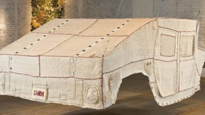 A large, suspended sculpture resembling a military vehicle is crocheted from beige yarn, displayed in an art gallery with white brick walls and vehicle sketches. [gen-ai]