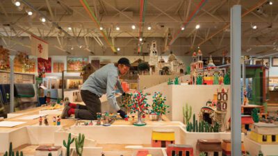 A person kneels on a display platform arranging miniature figurines and buildings in a brightly lit museum with colorful folk art exhibits. [gen-ai]