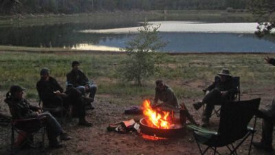 A group of people sit around a campfire near a lake at dusk, surrounded by trees and outdoor chairs. [gen-ai]