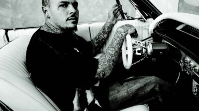 Tattooed man with a shaved head sits in the driver’s seat of a classic convertible car, holding the steering wheel and looking toward the camera. [gen-ai]