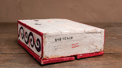 A rectangular, hand-painted white and red brick sits on a wooden surface. One side features a black swirl pattern and numbers are printed upside-down on the side facing up. [gen-ai]