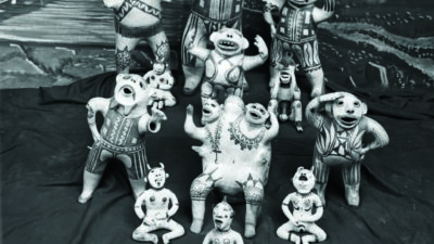 A group of decorated pottery figurines arranged on a cloth, labeled as Pottery figures, By Pueblos of New Mex. with a painted backdrop behind them. [gen-ai]