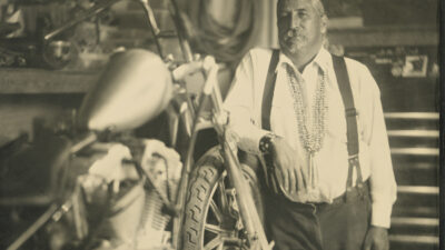A man in suspenders stands beside a motorcycle in a workshop, surrounded by tools and equipment, with a neutral expression. [gen-ai]