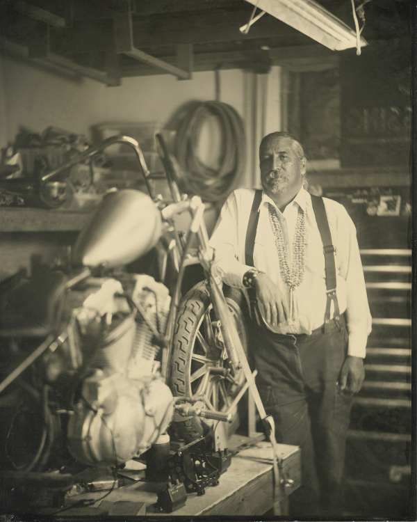 A man in suspenders stands beside a motorcycle in a workshop, surrounded by tools and equipment, with a neutral expression. [gen-ai]