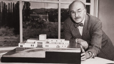 A man wearing glasses and a bow tie stands beside a scale model of a modern building, with large windows and a landscape visible in the background. [gen-ai]