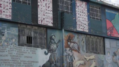 A two-story building with murals and posters on the exterior walls, depicting faces, text, and various human and animal figures. The windows have metal bars. [gen-ai]