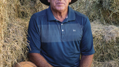 A middle-aged man in a cowboy hat and blue shirt sits on stacked hay bales, looking directly at the camera. [gen-ai]