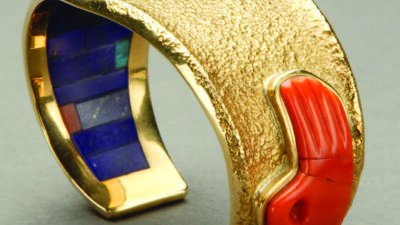 A wide, textured gold cuff bracelet with an inlaid red stone shaped like a hand and blue and green inlays inside the band. [gen-ai]