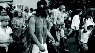 A group of people in bohemian attire walk together outdoors, some holding hands and smiling, in a black-and-white photo. [gen-ai]
