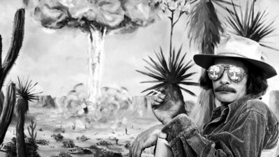 A man in a hat and sunglasses sits in a chair, smoking, with a desert landscape and a large nuclear explosion painted on the wall behind him. [gen-ai]