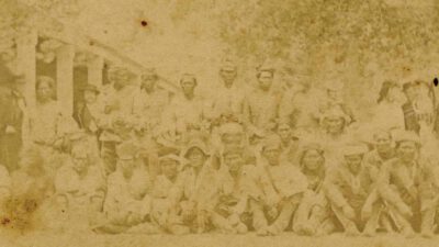 A sepia-toned, faded photograph of a large group of people outdoors, posed in rows under trees, with most sitting and some standing. [gen-ai]