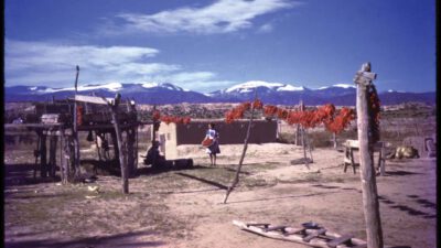 Adobe home with hanging red chilies, wooden structures, and a person standing outside; snow-capped mountains and clear sky in the background. [gen-ai]