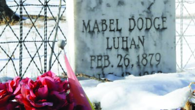 A gravestone for Mabel Dodge Luhan, dated February 26, 1879, with flowers and rocks placed on top, sits in a snowy outdoor setting. [gen-ai]