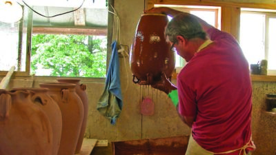 A man in a workshop dips a large clay pot into a container of glaze, with other unfinished pots on a nearby table. [gen-ai]