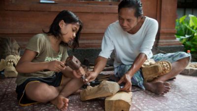 An adult and a child sit on a tiled floor, working together on wood carving using chisels and mallets, with carved pieces and tools around them. [gen-ai]