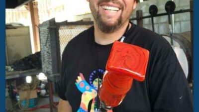 A smiling man wearing sunglasses and a black shirt holds a red glassblowing piece in a workshop. Text reads: Encounter Culture, Season 1 Episode 4. [gen-ai]