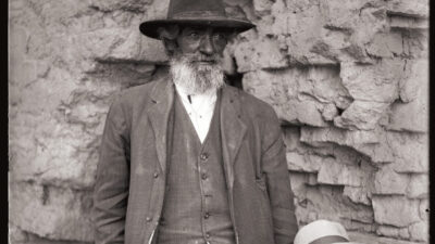 An older man and a young boy wearing hats and suits stand in front of a rough stone wall, looking directly at the camera. [gen-ai]