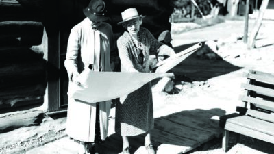 Two elderly women in coats and hats stand outside on a wooden path, looking at and discussing a large rolled-out paper or blueprint in front of a log building. [gen-ai]