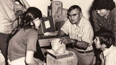 A man demonstrates stone carving to four young adults gathered around him, observing closely in what appears to be an art classroom or workshop. [gen-ai]