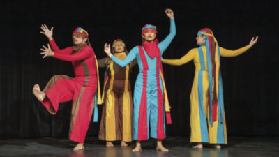Four dancers on a stage with a black background in various poses, several with arms extended wide.