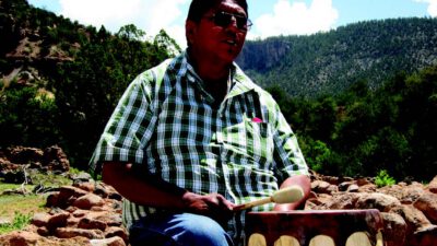 A person wearing sunglasses and a plaid shirt plays a hand drum outdoors, surrounded by rocks and trees with mountains in the background. [gen-ai]