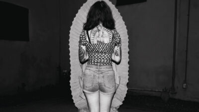 A person with tattoos stands facing a scalloped-edged backdrop, seen from behind, wearing a polka-dot top and denim shorts in a dimly lit setting. [gen-ai]